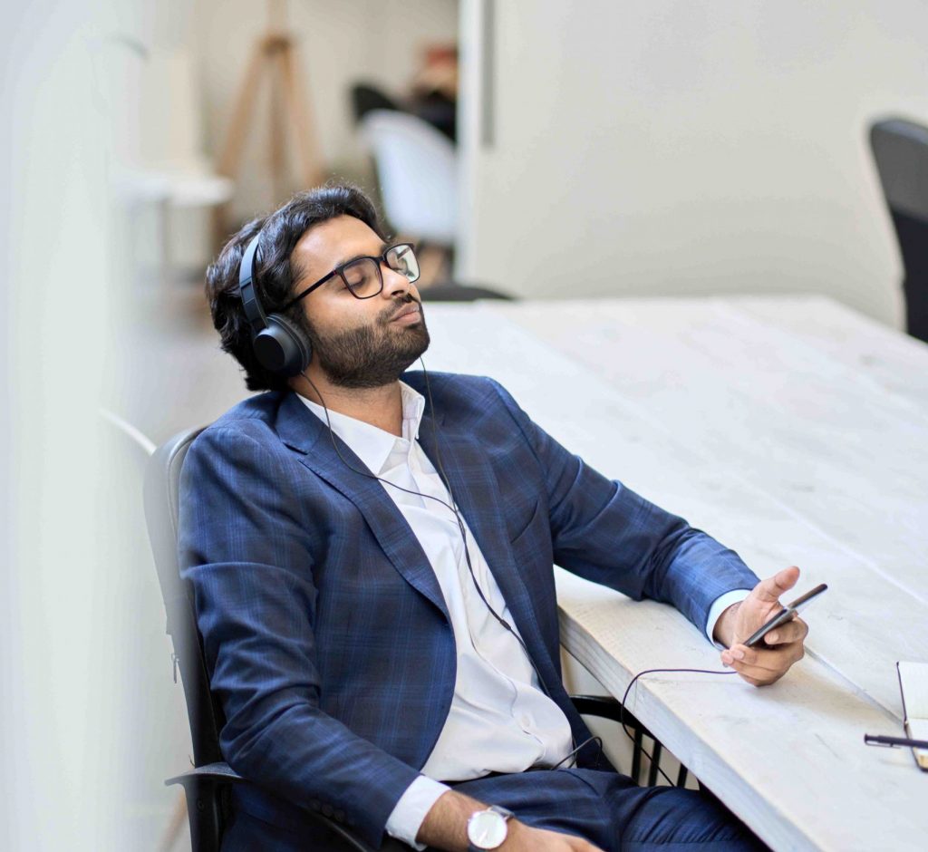 Indian business man employee executive wearing suit and headphones sitting at work desk using phone listening to mobile music, podcast, enjoying audio book taking break after work. Vertical